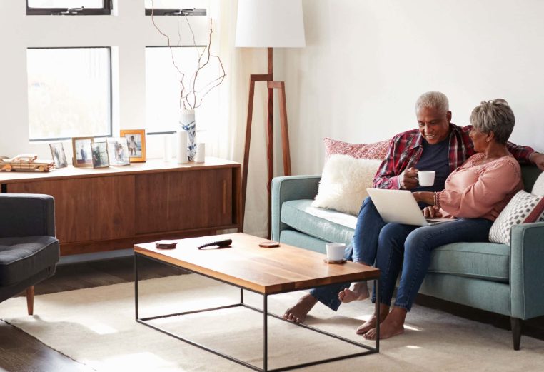 An older couple sit on a living room couch looking together at a laptop.