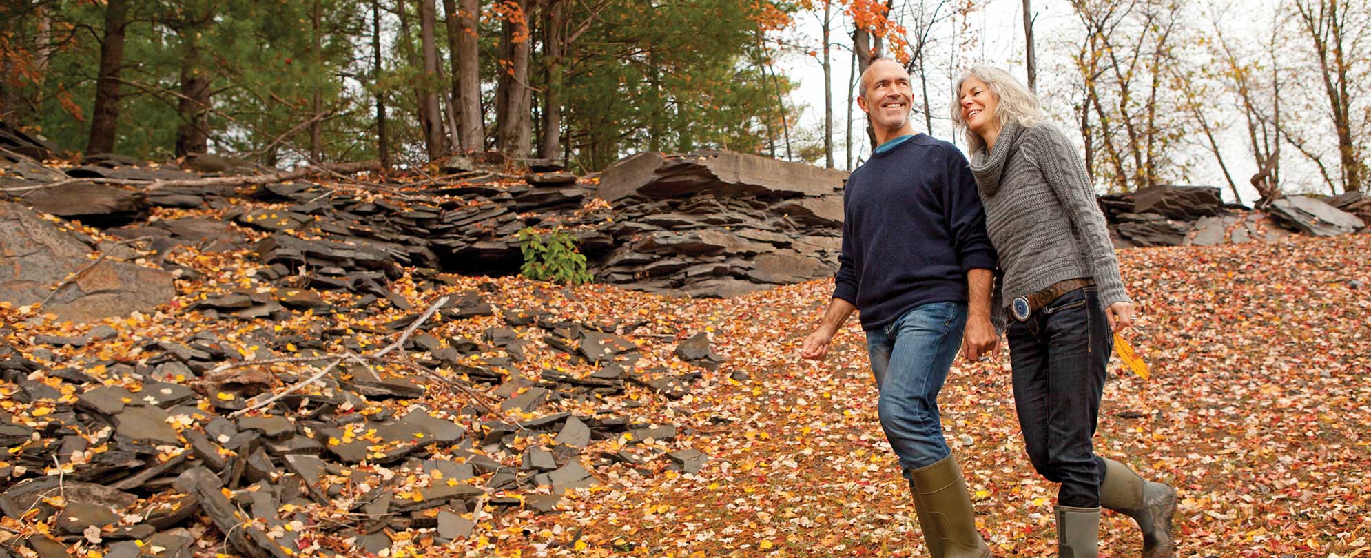 An older couple walks holding hands on a rocky mountain trail covered in fall leaves.