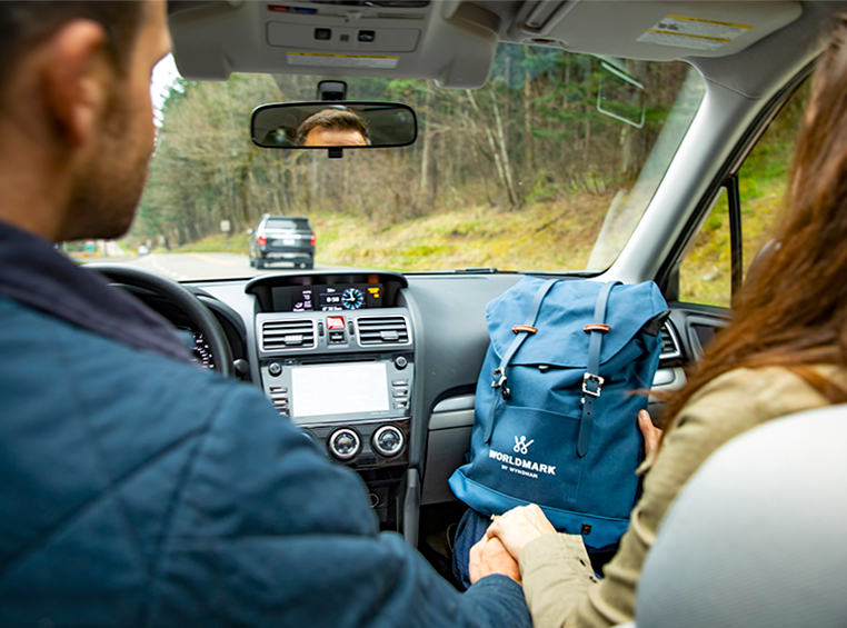 A man and woman in the front seats of a car, the woman is holding a WorldMark by Wyndham backpack on her lap.