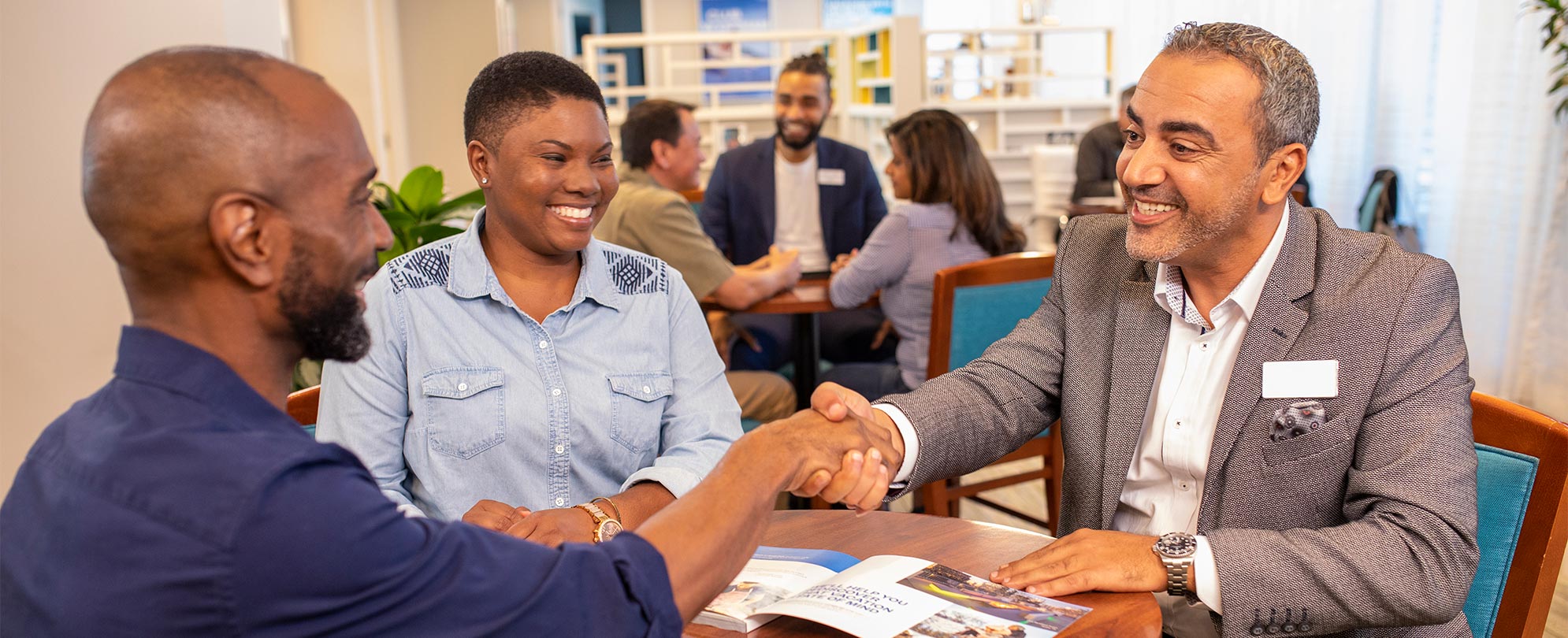 A WorldMark by Wyndham associate sits at a table with a smiling man and woman, he shakes the man’s hand.