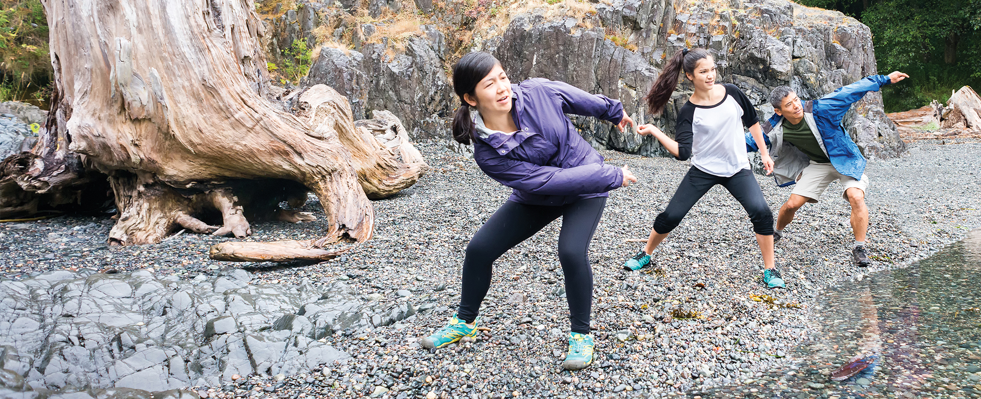 A mom, dad, and teenage daughter stand on a rocky shoreline focus as they throw stones into the water.