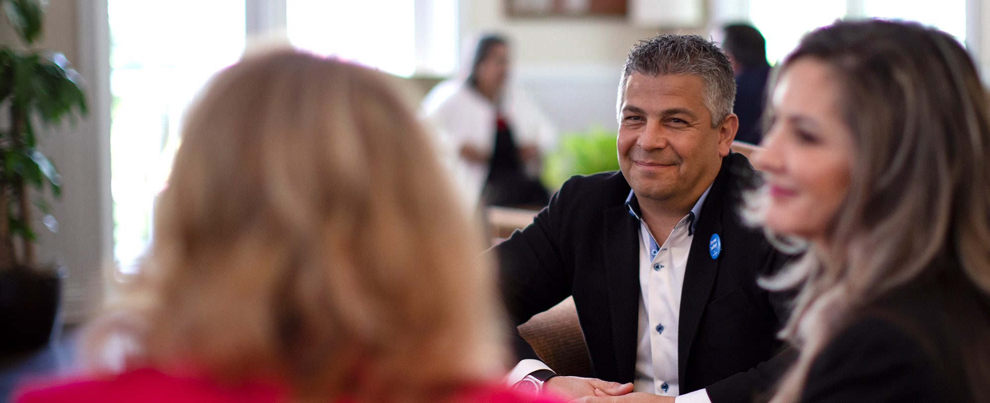 Two people are seated at a table looking and smiling at the person speaking at the head of the table.