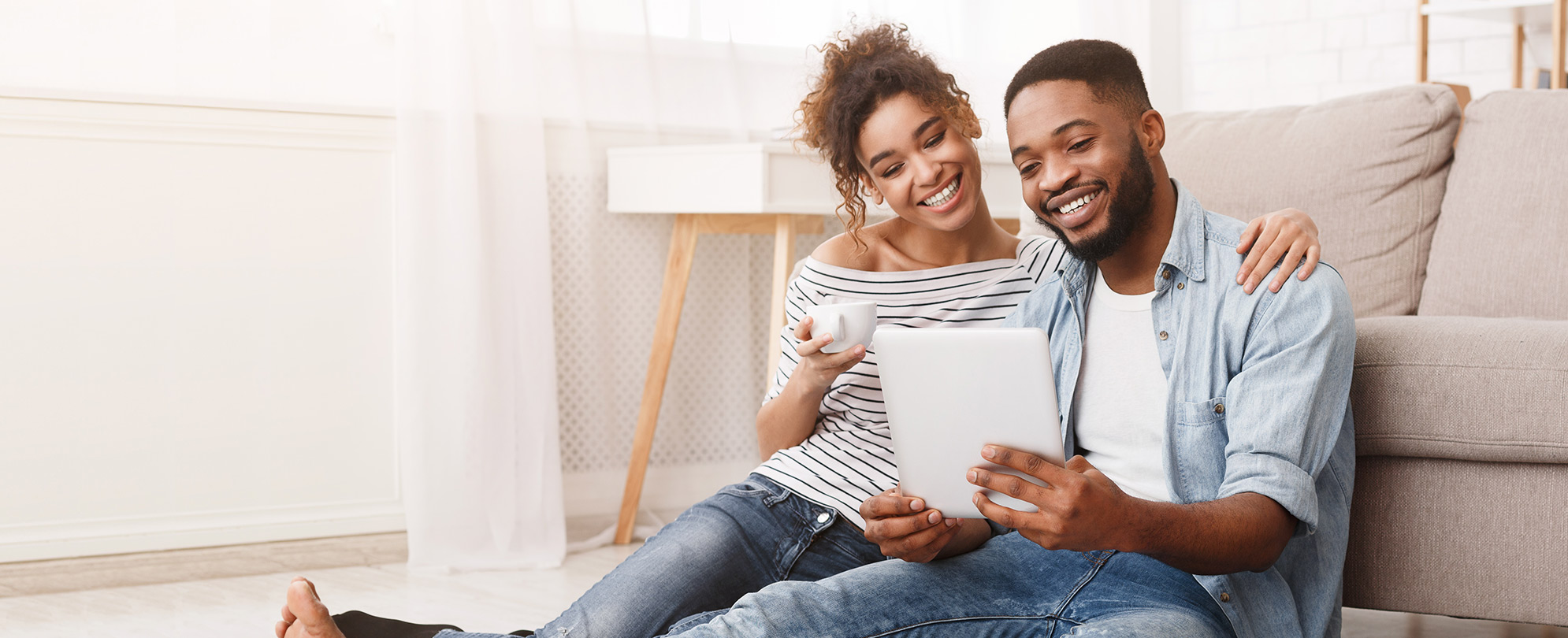 A couple sits comfortably on the floor in front of a couch, smiling down at a tablet.