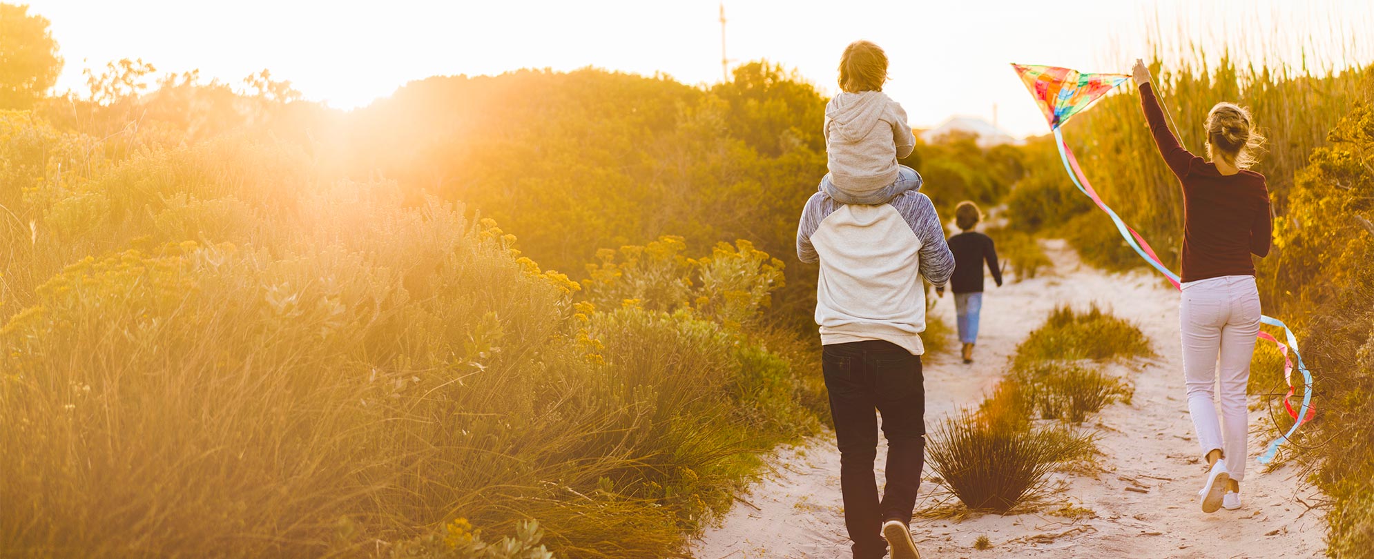 A family walks along a sandy trail, the dad carries their young child on his shoulders while the mom flies a kite.