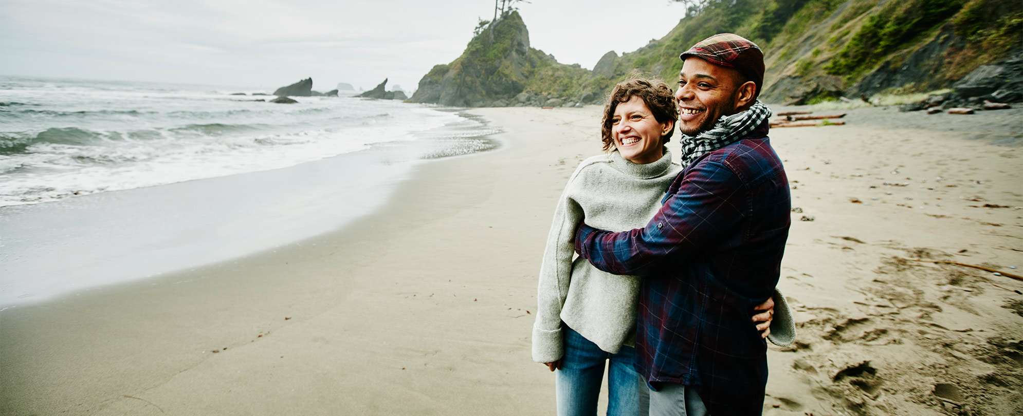 A man and woman wearing winter clothes stand embracing and smiling on a beach, with lush, rocky mountains in the background.