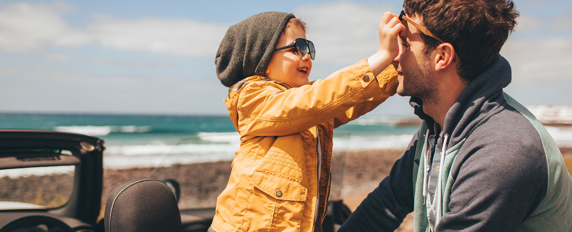 A dad and son sit in a roofless car on a beach, the young boy reaches to adjust his dad's sunglasses.