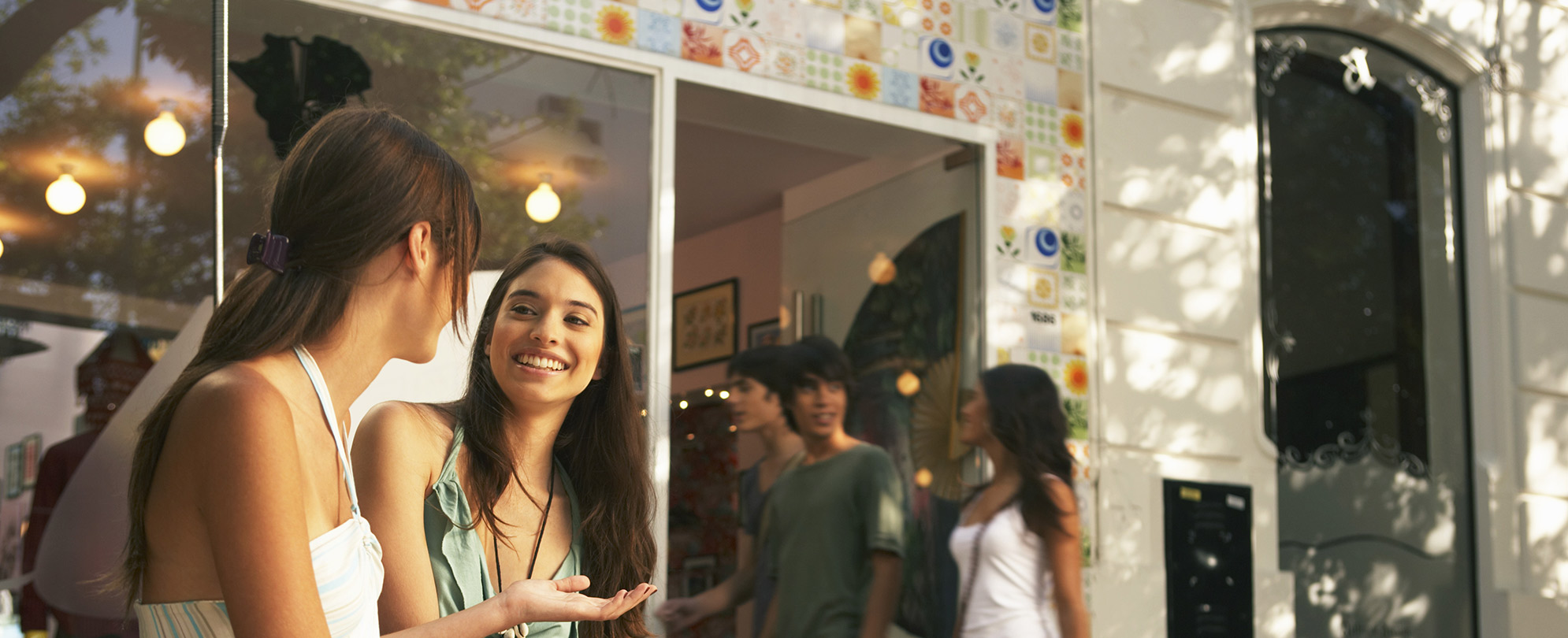 Two women chat outside as a small group of people walks through a nearby doorway surrounded by multicolored tiles.