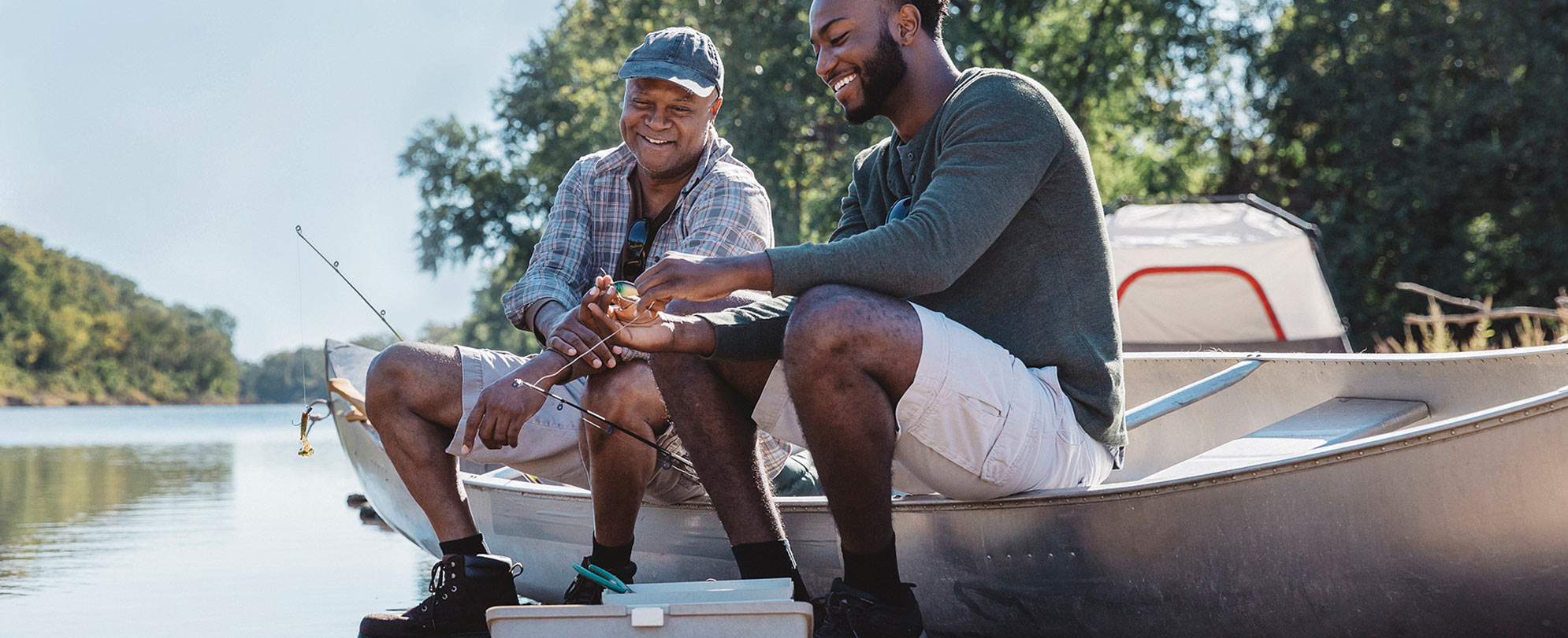 A dad and his adult son smile as they sit on a canoe by the shore of a lake, putting bait on a fishing line. 