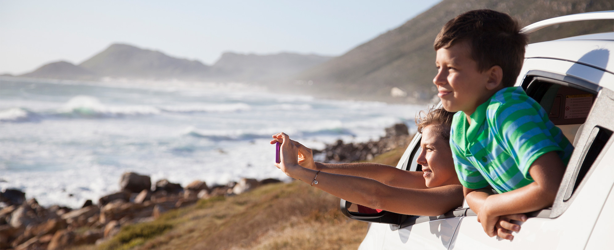 Two children lean out of car window while taking a photograph of beautiful beach scenery.