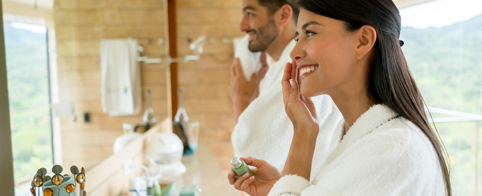 A man and woman both wearing hotel robes getting ready for their day. The woman is smiling, putting cream on her face, and the man is standing smiling at the mirror. 